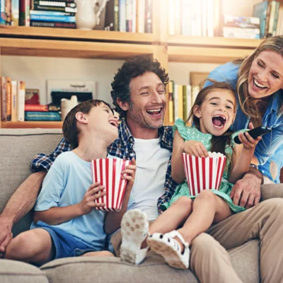 Foto: Glückliche Familie (Sohn, Mann, Tochter, Frau) auf grauer Couch vor hölzernen Bücherregal