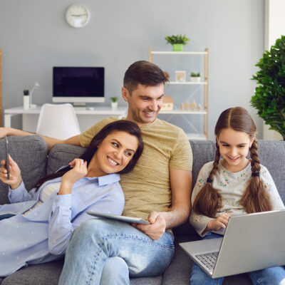 Foto: Glückliche Familie nutzt einen Laptop zu Hause; Eltern und Mädchen schauen auf dem Sofa sitzend ein Video